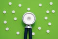 The medical stethoscope is on a green background surrounded by white round pills. The concept of a minimalistic photo to indicate