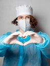 Medical worker in a disposable mask and a lab coat makes a heart sign. Gray homogeneous background. Conceptual photo: doctors on