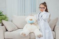 Pretty little girl wearing doctor coat, stethoscope and glasses, standing, holding hand on the head of teddy-bear in Royalty Free Stock Photo