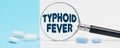 On a blue and white background, there are pills and a magnifying glass, inside of which it is written - TYPHOID FEVER