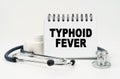 On a white table lies a stethoscope, pills and a notebook with the inscription - TYPHOID FEVER