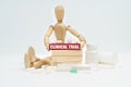 A figurine of a man among the pills, lifts a red wooden block with the inscription CLINICAL TRIAL from the block wall