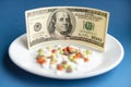 Medicines costs money. Drugs and dollars.White plate with multi-colored pills and capsules and a 100 dollar bill on a Royalty Free Stock Photo