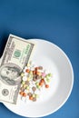 Medicines costs money. Drugs and dollars.White plate with multi-colored pills and capsules and a 100 dollar bill on a Royalty Free Stock Photo