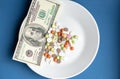 Medicines costs money. Drugs and dollars.White plate with multi-colored pills and capsules and a 100 dollar bill on a Royalty Free Stock Photo