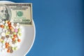 Medicines costs money. Drugs and dollars.White plate with multi-colored pills and capsules and a 100 dollar bill on a Royalty Free Stock Photo