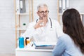 Serious old man doctor in white coat and glasses shows bottle to lady patient at table with jars of pills