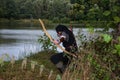 Medieval archer with black hood kneels on the ground before a lake, aims with arrow and curve forwards