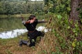 Medieval archer with black hood kneels at the side on the ground before a lake, aims with arrow and curve aside