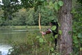 Medieval archer with black hood stands hidden behind tree in the lake with tense curve