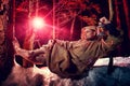 Medieval monk in canvas sackcloth robe with lattern resting on snow in dark forest and red light on winter night. Fantasy or fairy Royalty Free Stock Photo