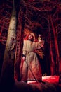 Medieval monk in canvas sackcloth robe with sword and lattern in dark forest and red light of moon on winter night. Fantasy or Royalty Free Stock Photo