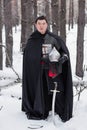 Medieval warrior in armor, helmet, black cloak with a saber in his hands walking in the winter in the forest Royalty Free Stock Photo