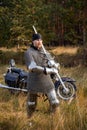 medieval warrior in armor with a two-handed sword in his hands stands next to a motorcycle against the backdrop of a forest