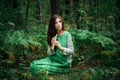 A medieval woman in a green dress plays on a wooden flute sitting in fern bushes. A girl in a gloomy forest plays music on a handm