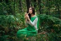 A medieval woman in a green dress plays on a wooden flute sitting in fern bushes. A girl in a gloomy forest plays music on a handm