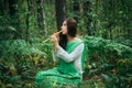 A medieval woman in a green dress plays on a wooden flute sitting in fern bushes. A girl in a gloomy forest plays music on a handm