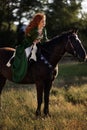 Medieval woman princess in green dress sits astride black steed horse.