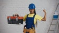 Medium shot of a dark-skinned young female construction worker standing in the room, wearing a tool belt, raising a tool Royalty Free Stock Photo