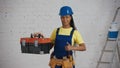 Medium shot of a dark-skinned young female construction worker standing in the room, wearing a tool belt, showing a tool Royalty Free Stock Photo