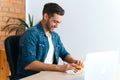 Medium shot of hungry happy freelancer male unwraping hamburger with beef from fast food restaurant sitting at working