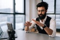 Medium shot of Indian male blogger in glasses broadcasting from office, using smartphone and tripod, sitting at table Royalty Free Stock Photo