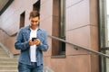 Medium shot portrait of cheerful handsome young man using mobile phone standing on stairs of modern office building. Royalty Free Stock Photo