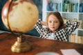 Medium shot portrait of curious child girl sitting on chair with hands behind head and looking at little globe on table Royalty Free Stock Photo