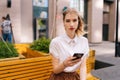 Medium shot of serious pretty young woman sitting on yellow bench on city street and looking at camera holding mobile Royalty Free Stock Photo