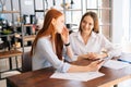 Medium shot of two happy young business women working using digital tablet at meeting desk with job documents at office. Royalty Free Stock Photo