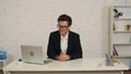 Medium shot of a young businessman wearing glasses, sitting at the table in an office, working on his laptop, focused