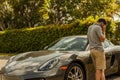 Medium shot of a young man at the phone, standing close to a Porsche Cayman. Urban scene.