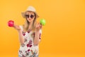 Medium studio shot of a confused caucasian blonde girl in a floral blouse, sunglasses, and a hat holding two apples -