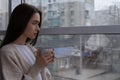 Melancholic young woman with drink looking out of window on rainy day, space for text. Loneliness concept Royalty Free Stock Photo