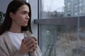 Melancholic young woman with drink looking out of window on rainy day, space for text. Loneliness concept Royalty Free Stock Photo