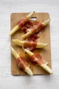 Melon slices wrapped in prosciutto on bamboo board over white wooden background, top view. Closeup. From above Royalty Free Stock Photo