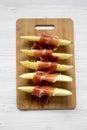 Melon slices wrapped in prosciutto on bamboo board over white wooden surface, top view. Close-up. From above Royalty Free Stock Photo