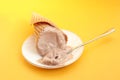 Melted chocolate ice cream in a waffle cone on a white saucer with a teaspoon, yellow background Royalty Free Stock Photo