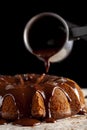 Melted hot chocolate pudding is being poured down from a metal coffee pot pitcher onto a chocolate cake as icing.