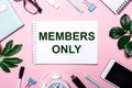 MEMBERS ONLY is written in a white notebook on a pink background surrounded by business accessories and green leaves