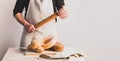 A men baker in a cotton apron holds a wooden rolling pin. Various loaves of fresh bread on the kitchen table.