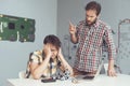 A man chastises the teenager who broke the robot. A teenager is sitting at a table with his head in his hands