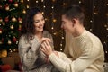 The man gives the girl an engagement ring, couple is in christmas lights and decoration, dressed in white, fir tree on dark wooden Royalty Free Stock Photo