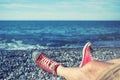 Men`s legs in shorts and red sneakers on a pebbly beach by the sea on a sunny day.