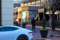 2 men standing and one man walking in a shopping center surrounded by buildings, cars and a black flower pot with colorful flowers