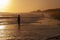 Men surf fishing among ocean waves at sunset with a power plant in the background