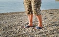 Men& x27;s feet in flip-flops and shorts, a man standing on the beach on a pebble beach, sunrise in the morning