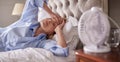 Menopausal Mature Woman Suffering With Insomnia In Bed At Home Using Electric Fan
