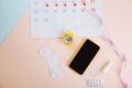 Menstruation calendar and smartphone with cotton tampon, sanitary pad and yellow alarm clock on blue pink background