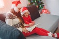 Merry Christamas and Happy New Year. Young woman with red hat sit on sofa with daughter. She holds book on her lap